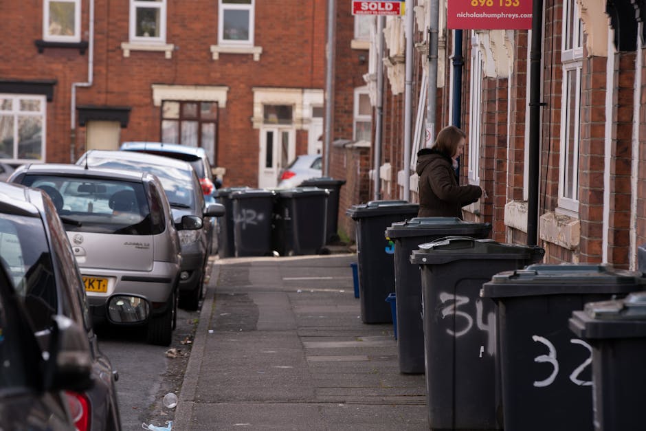 The image depicts a residential street with red brick terraced houses, where a row of black refuse bins is lined up along the pavement. The bins, made of plastic with lids, are positioned on the sidew